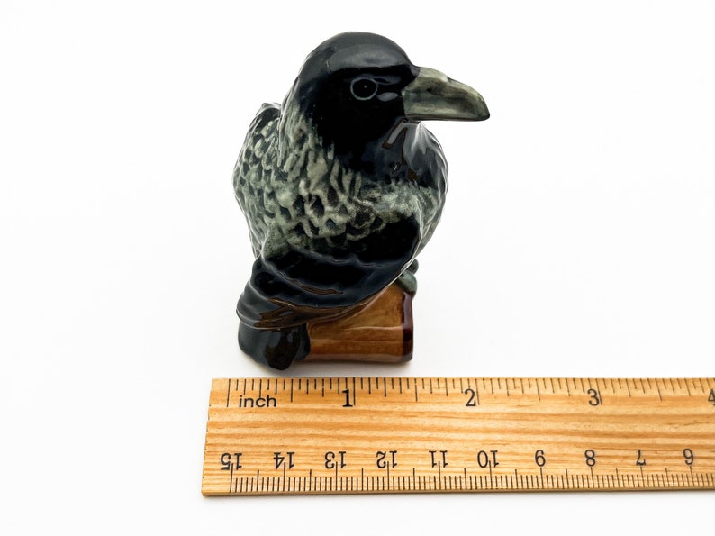 May include: A black and green ceramic crow figurine perched on a wooden base. The crow is about 2 inches tall.