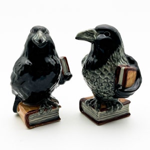 May include: Two black ceramic raven figurines holding books. The ravens are perched on stacks of books with brown covers.
