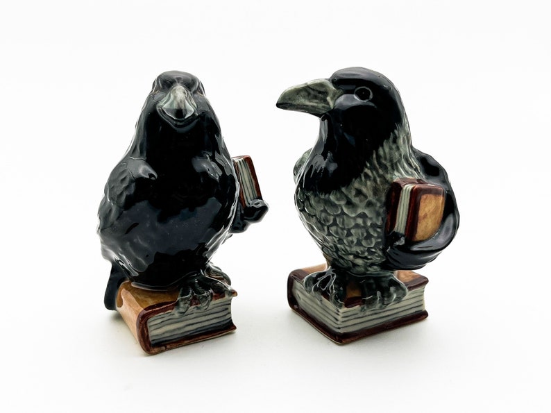 May include: Two black ceramic raven figurines holding books. The ravens are perched on stacks of books with brown covers.