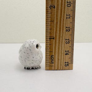 K&ouml;nnte beinhalten: Kleine, wei&szlig;e Keramik-Eulenfigur mit schwarzen Sprenkeln und goldenen Augen. Die Eule ist etwa 2,5 cm hoch. Ein Holzlineal liegt neben der Eule und zeigt das Ma&szlig; in Zoll an.