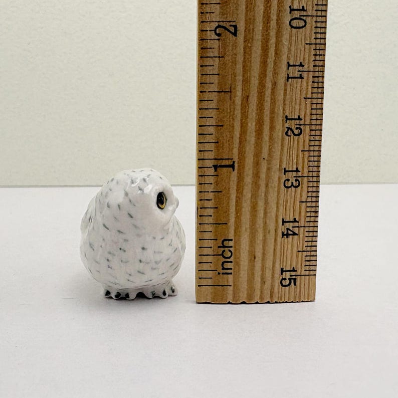 K&ouml;nnte beinhalten: Kleine, wei&szlig;e Keramik-Eulenfigur mit schwarzen Sprenkeln und goldenen Augen. Die Eule ist etwa 2,5 cm hoch. Ein Holzlineal liegt neben der Eule und zeigt das Ma&szlig; in Zoll an.