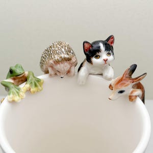 以下が含まれることがあります： 白いセラミック製のマグカップの縁に、カエル、ハリネズミ、猫、鹿の4つのセラミック製の動物の置物が置かれています。