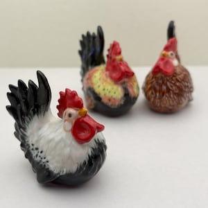 May include: Three ceramic rooster figurines. The roosters have red combs and wattles, with black, white, and brown plumage. The figurines are small and detailed, with a glossy finish. The roosters are arranged on a white surface.