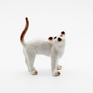 May include: A white porcelain figurine of a Siamese cat with brown markings on its tail and face. The cat is standing on all fours with its head tilted up.