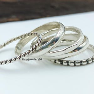 Könnte beinhalten: Eine Sammlung von Silberringen in verschiedenen Designs. Ein Ring hat ein gedrehtes Seilmuster, während andere glatte oder strukturierte Bänder aufweisen. Die Ringe werden auf einer weißen Oberfläche mit einem dunklen Holzelement im Hintergrund präsentiert.