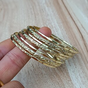 Könnte beinhalten: Ein Stapel goldfarbener Armreifen mit floralen und strukturierten Designs. Die Armreifen sind gestapelt und reflektieren das Licht. Der Schmuck liegt auf einer hellen Holzoberfläche.