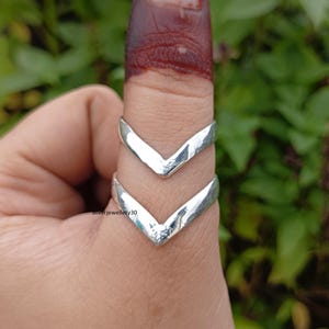 Könnte beinhalten: Zwei silberne V-förmige Ringe werden an einem Finger getragen. Die Ringe haben eine gehämmerten Textur und sind übereinander positioniert. Der Hintergrund ist eine verschwommene Ansicht von grünem Laub.