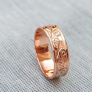 Pu&ograve; includere: Un anello in oro rosa con un motivo floreale inciso. L'anello ha una fascia con un disegno ripetuto di foglie e volute. L'anello &egrave; mostrato su una superficie grigio chiaro.