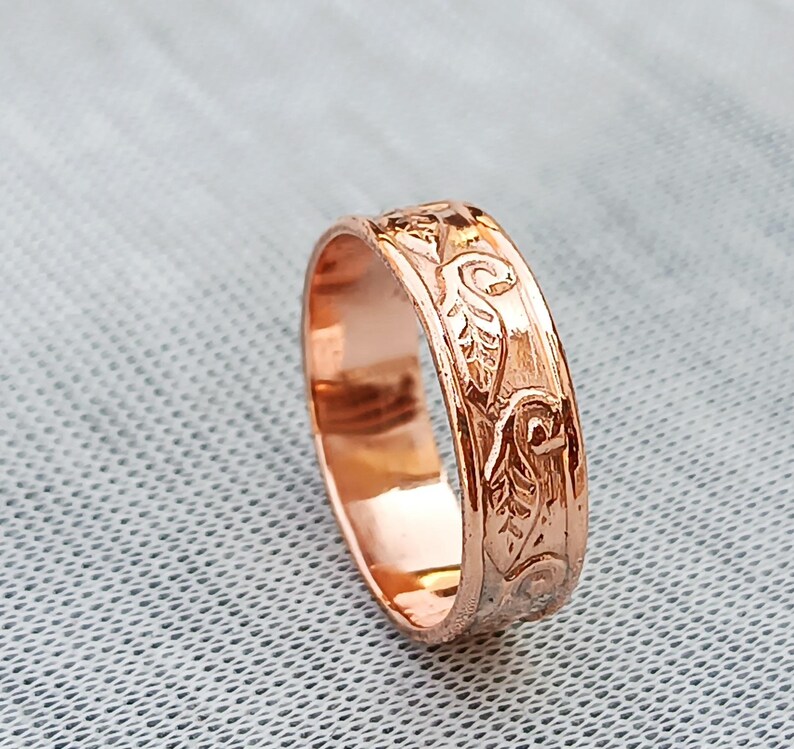 Pu&ograve; includere: Un anello in oro rosa con un motivo floreale inciso. L'anello ha una fascia con un disegno ripetuto di foglie e volute. L'anello &egrave; mostrato su una superficie grigio chiaro.