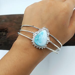 Könnte beinhalten: Silbernes Armreif-Armband mit einem hellblauen, birnenförmigen Edelstein. Der Edelstein ist in einem dekorativen Silberrahmen mit strukturiertem Rand gefasst. Das Armband hat drei parallele Silberbänder.