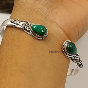 Könnte beinhalten: Offener Armreif mit Silberfiligran und zwei tropfenförmigen Malachitsteinen. Die grünen Steine sind in verzierten Silberfassungen gefasst. Das Armband wird am Handgelenk getragen.