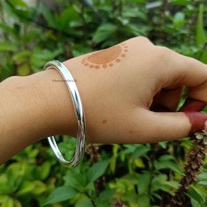 Könnte beinhalten: Ein glänzendes, silberfarbenes Armreif-Armband. Das glatte, abgerundete Design reflektiert das Licht und erzeugt ein helles Aussehen. Das Armband wird am Handgelenk getragen, mit einem verschwommenen grünen Hintergrund.