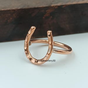 Könnte beinhalten: Ein Ring in Roségold-Optik mit einem hufeisenförmigen Mittelstück, das eine gehämmerte, strukturierte Oberfläche aufweist. Die Ringschiene ist glatt und kreisförmig und mit der Basis des Hufeisens verbunden. Der Text Silverjewellery30 ist sichtbar.
