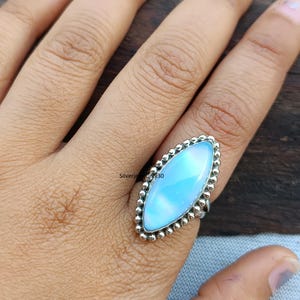 Könnte beinhalten: Ein Silberring mit einem großen, ovalen, hellblauen Edelstein. Der Edelstein ist von einem perlenbesetzten Silberrand umgeben. Der Ring wird auf einer Hand vor einem Holzgrund gezeigt. Der Text "Silverjewelry30" ist auf dem Ring sichtbar.