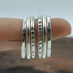 Könnte beinhalten: Eine Ansammlung von Silberringen, jeder mit einem einzigartigen strukturierten Design, ist auf einem Finger aufgereiht. Die Ringe variieren in der Textur, einige haben gehämmerten oder geriffelten Oberflächen. Der Hintergrund ist unscharf.