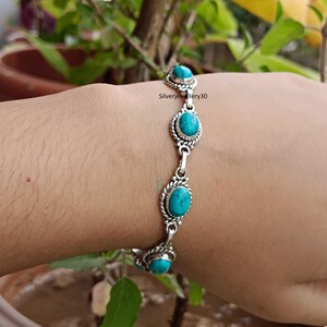 Könnte beinhalten: Ein silbernes Armband mit ovalen Türkissteinen. Das Armband hat ein detailliertes Design mit einem geflochtenen Rand um jeden Stein. Die Türkissteine haben eine leuchtende blaugrüne Farbe. Das Armband wird am Handgelenk getragen.