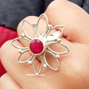 Könnte beinhalten: Ein silberner Ring mit einem roten Edelstein in der Mitte, der die Form einer Blume mit sechs Blütenblättern hat. Der Ring wird an einem Finger getragen.