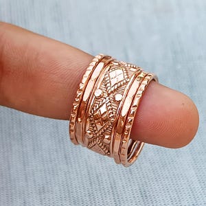 Könnte beinhalten: Ein Stapel kupferfarbener Ringe mit aufwendigen geometrischen Mustern und Texturen. Die Ringe variieren in der Breite und im Design, einige weisen Diamanten- und Punktmotive auf. Die Ringe sind auf einem Finger gestapelt, wodurch die Details des Schmucks hervorgehoben werden.