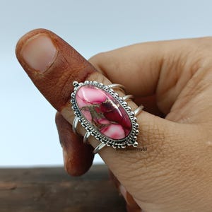 May include: A sterling silver ring featuring an oval-shaped pink and maroon gemstone. The ring has a detailed, ornate setting with a textured border. The ring is on a finger, showcasing the design and craftsmanship.
