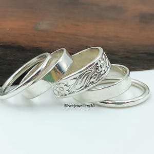 Könnte beinhalten: Eine Sammlung von Silberringen, darunter ein dünner Bandring, ein breiterer Ring mit floralen Details und ein glatter Ring. Die Ringe liegen auf einer weißen Oberfläche mit Holzgrund.