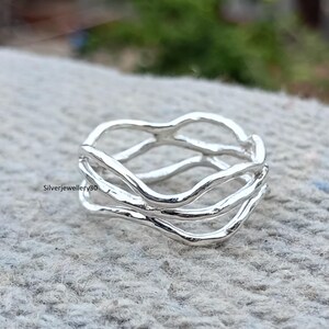 Può includere: Un anello d'argento dal design unico e fluido. L'anello presenta più fasce ondulate che si intrecciano per creare un aspetto organico e strutturato. L'anello è realizzato in metallo lucido e riflettente, e il design è intricato e accattivante.