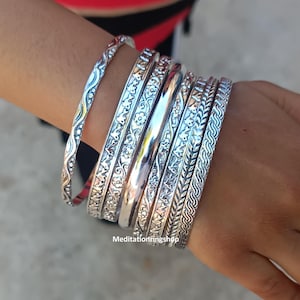 May include: A set of silver bangles with various intricate designs, including floral patterns, wavy lines, and braided textures.