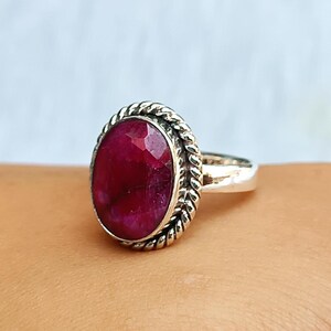 Könnte beinhalten: Ein Silberring mit einem ovalen, facettierten, tiefroten Edelstein. Der Edelstein ist in einem dekorativen, seilartigen Rand gefasst. Der Ring hat ein schlichtes, poliertes Silberband.