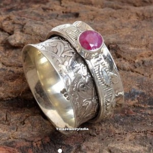 Könnte beinhalten: Ein silberner Drehring mit einem filigranen Blumenmuster und einem rosafarbenen Edelstein. Der Ring besteht aus zwei Bändern, die sich unabhängig voneinander drehen.