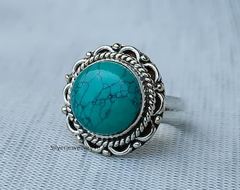 Può includere: Anello d'argento con una grande pietra turchese rotonda. La pietra è incastonata in una lunetta decorativa con un bordo a forma di corda e piccoli accenti dorati. L'anello ha una fascia semplice e lucida.