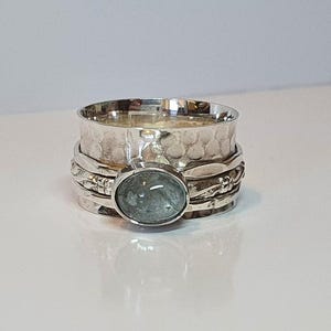 Könnte beinhalten: Ein silberner Spinner-Ring mit einem ovalen, hellblauen Edelstein. Der Ring hat ein strukturiertes Band und dekorative Details. Der Edelstein ist in einer Lünettenfassung gefasst. Der Ring wird auf einer weißen Oberfläche präsentiert.