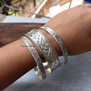 Könnte beinhalten: Vier silberne Armreifen mit Blumen- und Kleeblattmustern. Die Armreifen variieren in der Breite und weisen detaillierte Designs auf. Das Bild zeigt die Armreifen an einem Arm, wodurch die aufwendige Handwerkskunst und die reflektierende Qualität des Metalls hervorgehoben werden.