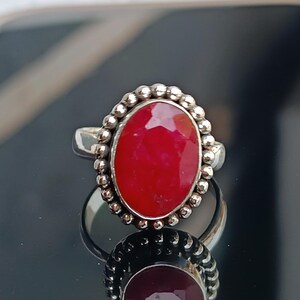 Könnte beinhalten: Ein silberner Ring mit einem großen, oval geschliffenen, leuchtend roten Edelstein. Der Edelstein ist von einem silbernen Perlenrand umgeben. Der Ring wird auf einer reflektierenden Oberfläche gezeigt.