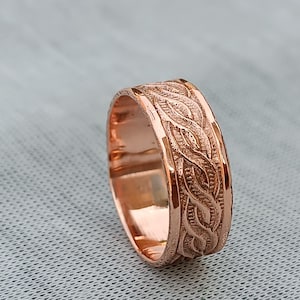 May include: Rose gold ring with a detailed, engraved pattern of intertwined leaves. The ring has a polished finish and a wide band, showcasing the intricate design. The ring is a piece of jewelry.