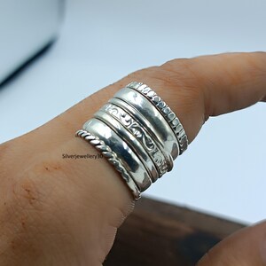 Könnte beinhalten: Mehrere silberne Ringe, die auf einem Finger getragen werden. Die Ringe weisen verschiedene Designs auf, darunter ein gedrehtes Seil, geätzte Muster und Perlenränder. Die Ringe sind glänzend und reflektieren das Licht. Die Ringe sind gestapelt und bilden ein auffälliges Schmuckstück.