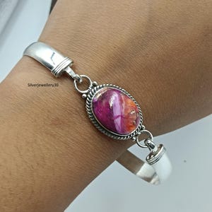 Könnte beinhalten: Silberarmband mit einem ovalen, mehrfarbigen Edelstein. Der Edelstein hat Schattierungen von Rosa, Lila und Orange. Das Armband hat ein schlichtes, elegantes Design mit einem strukturierten Rand um den Edelstein.