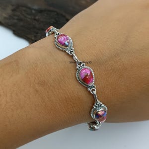 Könnte beinhalten: Ein silbernes Armband mit tropfenförmigen rosa und roten Steinen. Das Armband ist durch silberne Glieder verbunden und wird am Handgelenk getragen. Die Steine haben ein einzigartiges Muster.