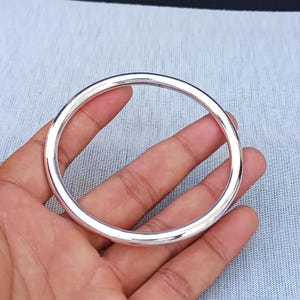 Op de afbeelding: Een glanzende, zilverkleurige armband. De ronde armband heeft een glad oppervlak en lijkt van metaal te zijn. De armband wordt in een hand gehouden tegen een lichtblauwe achtergrond.