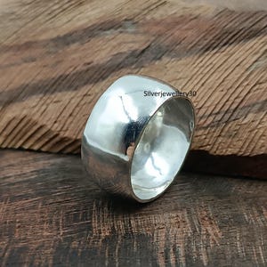 Könnte beinhalten: Ein breiter, polierter Silberring mit glattem, abgerundetem Design. Der Ring reflektiert das Licht und erzeugt ein helles, glänzendes Aussehen. Der Text "Silverjewellery30" ist auf dem Ring sichtbar. Der Ring befindet sich auf einem Holzgrund.