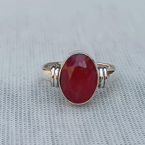 Könnte beinhalten: Ein silberner Ring mit einem großen, oval geschliffenen, tiefroten Edelstein. Der Edelstein ist der Mittelpunkt, gefasst in einem polierten Silberband mit dekorativen Seitenakzenten. Der Ring wird auf einer strukturierten, hellfarbigen Oberfläche präsentiert.