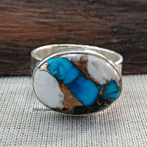 Può includere: Un anello d'argento con una pietra ovale. La pietra presenta un mosaico di colori bianco, turchese e oro. L'anello è posto su uno sfondo strutturato. Il testo "Silverjewellery" è visibile sull'anello.
