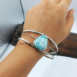 Könnte beinhalten: Ein silbernes Armreif-Armband mit einem großen, tropfenförmigen, hellblauen Edelstein. Das Armband hat drei parallele Bänder, mit einem gedrehten Drahtdetail zwischen den oberen beiden Bändern. Der Edelstein ist der Mittelpunkt des Armbands.