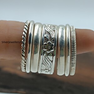 Könnte beinhalten: Eine Sammlung von Silberringen, jeder mit einem einzigartigen Design, auf einem Finger getragen. Die Ringe umfassen einen gedrehten Seilstil, glatte Bänder und einen floral gravierten Ring. Die Ringe sind aus glänzendem Silbermetall gefertigt.