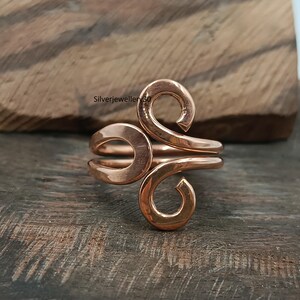 Könnte beinhalten: Ein polierter Kupferring mit einem Bypass-Design und zwei offenen, spiralförmigen Enden, die sich nach innen biegen. Die Metalloberfläche hat einen warmen, rötlich-braunen Farbton mit einer glatten, reflektierenden Oberfläche. Der Text Silverjewellery30 ist sichtbar.