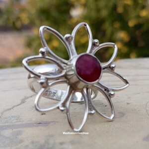 Puede incluir: Un anillo de plata con una piedra preciosa roja en el centro, con forma de flor de cinco p&eacute;talos. El anillo est&aacute; marcado con "925" en la banda.