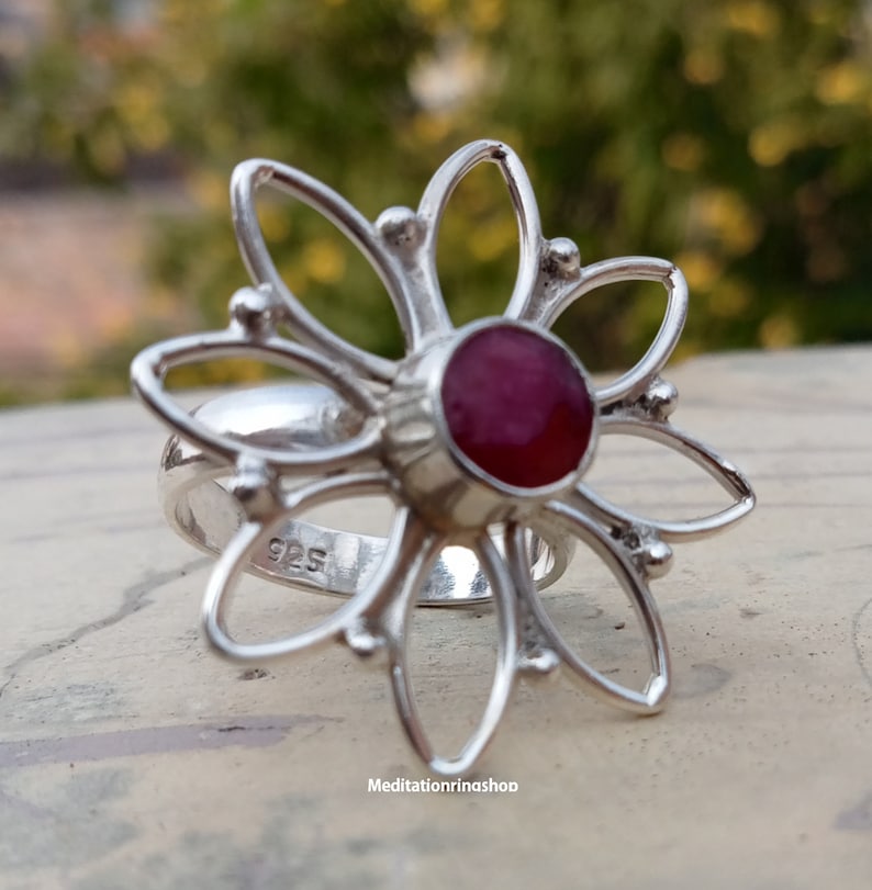 Puede incluir: Un anillo de plata con una piedra preciosa roja en el centro, con forma de flor de cinco p&eacute;talos. El anillo est&aacute; marcado con "925" en la banda.