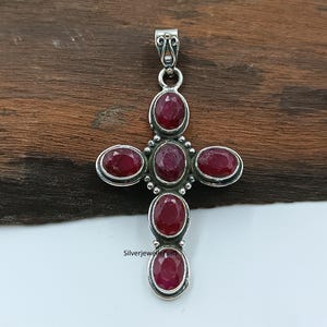 Könnte beinhalten: Silberfarbener Kreuzanhänger mit ovalen, tiefroten Edelsteinen. Das Kreuz hat eine dekorative Öse und ist vor einem Holzgrund platziert. Der Text "Silverjewellery36" ist sichtbar.