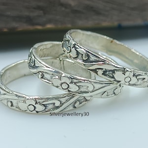 Könnte beinhalten: Drei silberne Ringe mit floralen Mustern. Jeder Ring zeigt ein sich wiederholendes Design aus Blumen und Blättern. Die Ringe sind gestapelt, wobei die floralen Details deutlich sichtbar sind. Die Ringe sind aus glänzendem Metall.