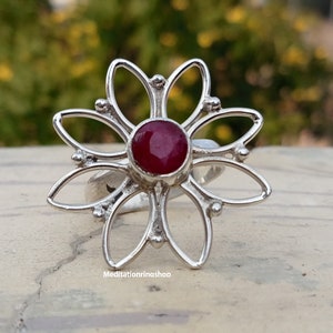 Puede incluir: Un anillo de plata con una piedra preciosa roja en el centro, con forma de flor de seis p&eacute;talos.