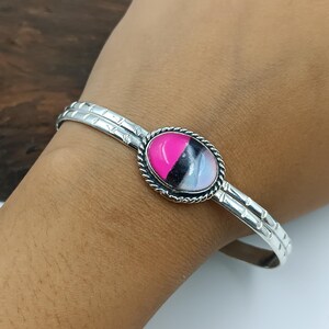 Könnte beinhalten: Ein silbernes Armreif-Armband mit einem ovalen Mittelstück. Das Mittelstück hat einen rosa, schwarzen und weißen Stein, der von einem gedrehten silbernen Seilmuster umrahmt wird. Das Armband hat ein strukturiertes Muster entlang des Bandes.