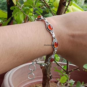 Könnte beinhalten: Ein silbernes Armband mit ovalen, rot-orangen Edelsteinen. Das Armband hat ein verziertes Design mit einer Reihe von Edelsteinen, die durch silberne Glieder verbunden sind. Der Text "Silverjewellery30" ist sichtbar.